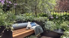 Garden bench with blanket, books and mug on it, surrounded by raised planters, verdant shrubs and trees