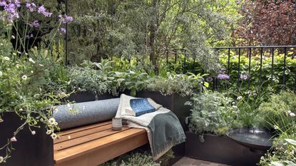 Garden bench with blanket, books and mug on it, surrounded by raised planters, verdant shrubs and trees