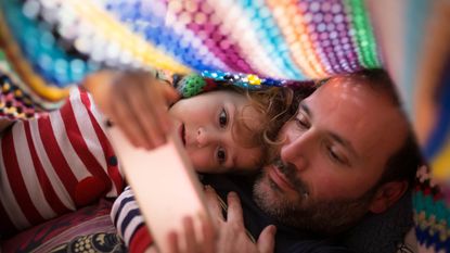 A dad and his little girl look at his phone under a blanket.