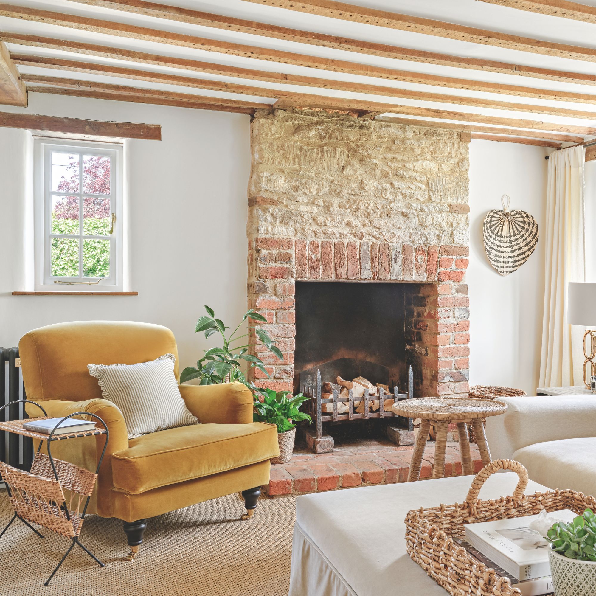 Cottage leaving room with wooden ceiling beams, white walls, large brick fireplace and statement yellow armchair. Large velvet pouffe in the center with basket and books on top