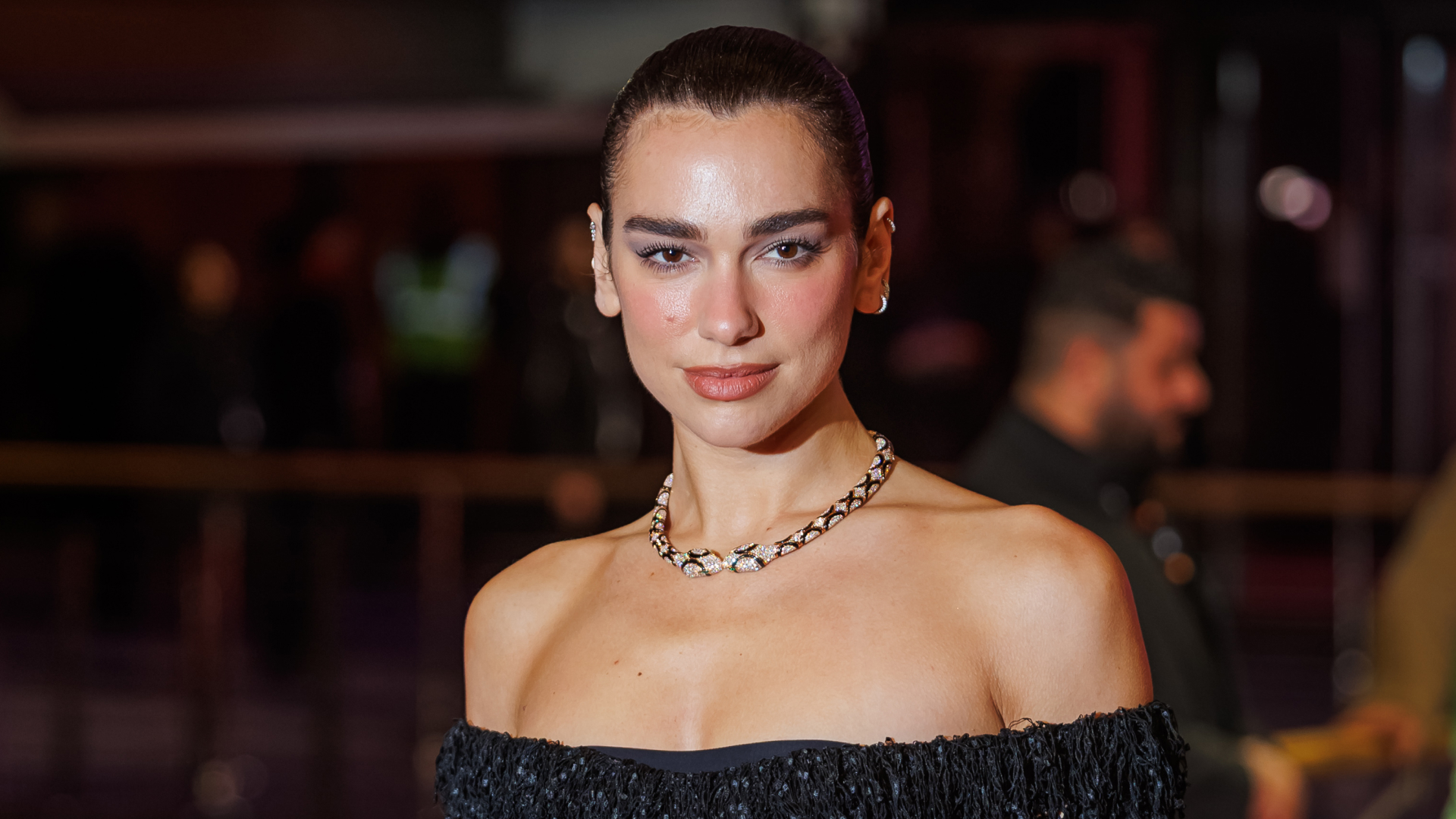 Dua Lipa wears an off-the-shoulder dress and wears her hair scraped back in a bun