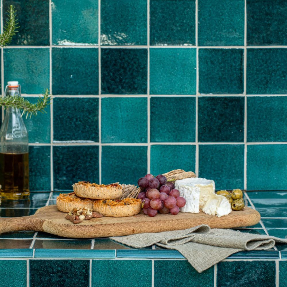 A teal glazed square tile kitchen splashback with a wooden cheeseboard, grapes, brie, olive oil and rosemary in the foreground.