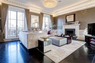 The living room includes grey tinted wallpaper, marble flooring, white fluffy sofas and carpet, large bifold doors which lead out onto the balcony with floor to ceiling curtains and art pieces around the room