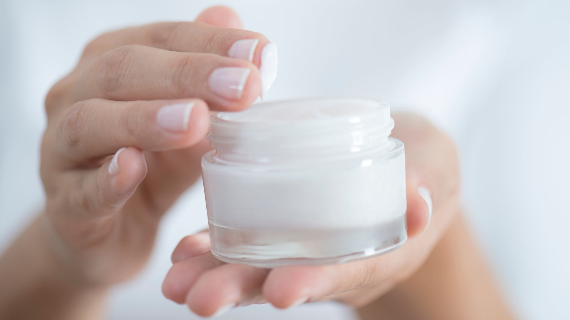 A woman's manicured hand dips into a glass jar of moisturizing lotion.