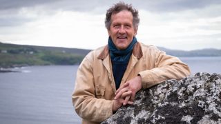 Monty standing by a big rock wearing a cream coat in front of the sea