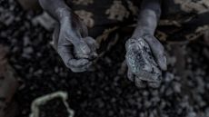 A pair of chalk covered hands hold small stones full of cobalt over a pile of blurry dark stones in the background.