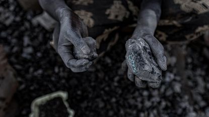 A pair of chalk covered hands hold small stones full of cobalt over a pile of blurry dark stones in the background.