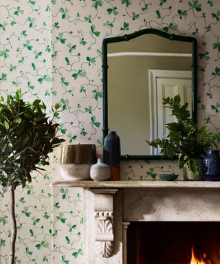 Green floral wallpaper, fireplace with green mirror placed above, houseplants, ornaments on mantel