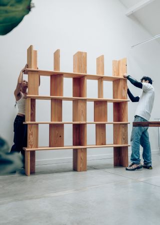 A woman and a man dressed in white tops and denim trousers work on the assembling of a wooden shelving unit.