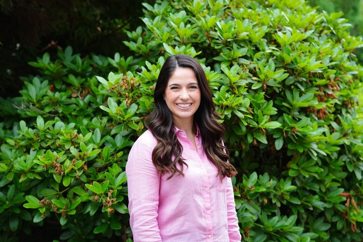Woman in pink shirt with dark brown hair against a large rhododendron bush
