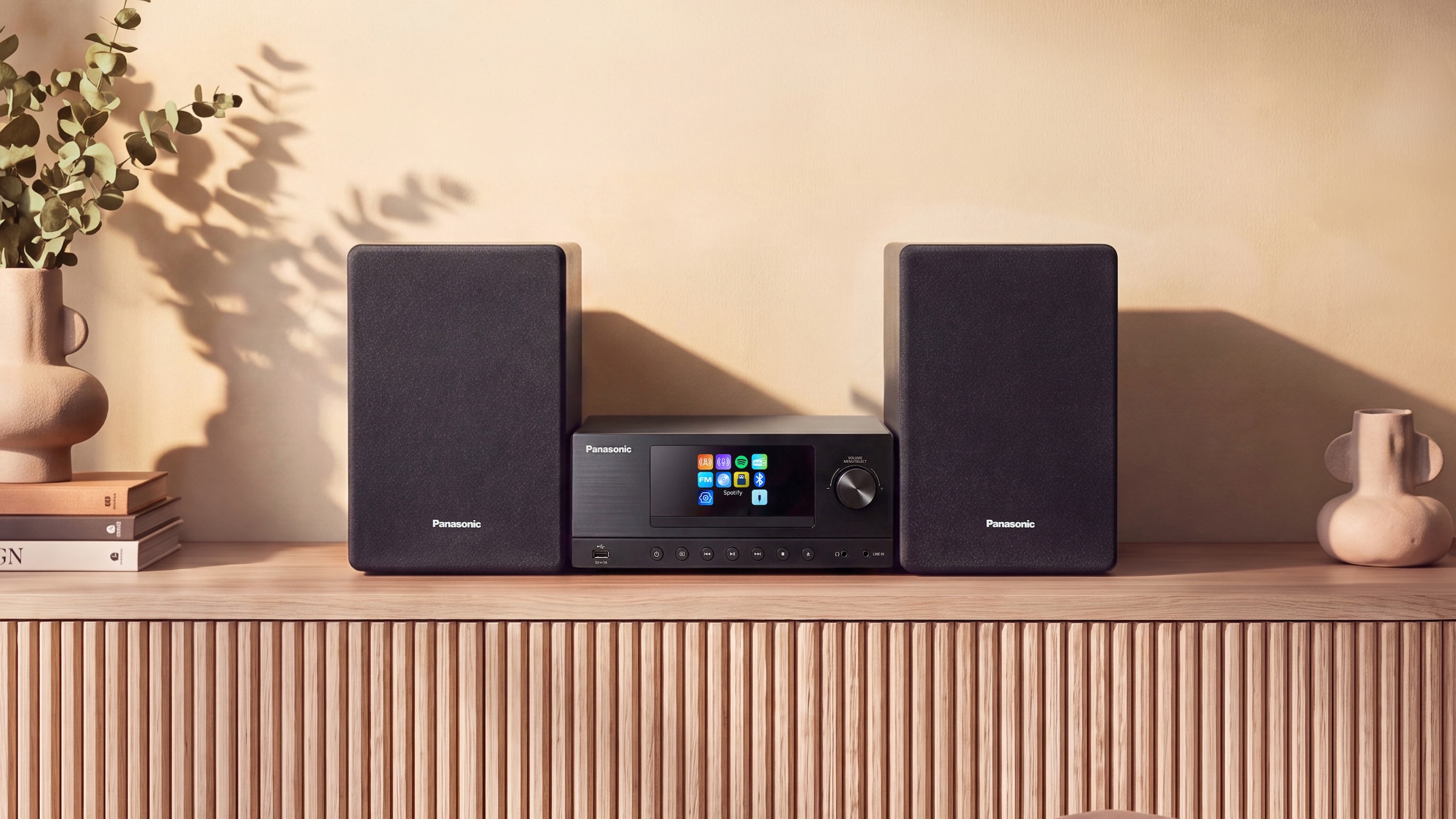 The Panasonic SC-PMX112 hi-fi system on a side unit next to a vase that sits on top of a stack of books.