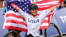 Paralympic handcyclist Oksana Masters holds up the US flag while wearing her white Team USA cycling kit during the 2024 Paris Paralympics.