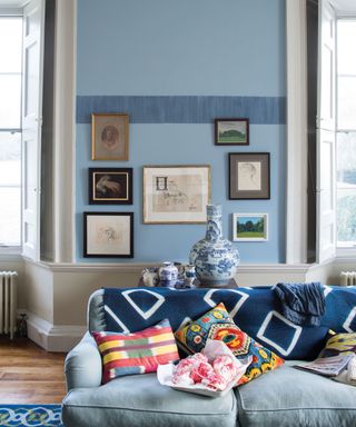 A blue living room with pale blue walls and a darker textured border, with white woodwork, a pale blue sofa and gallery wall.