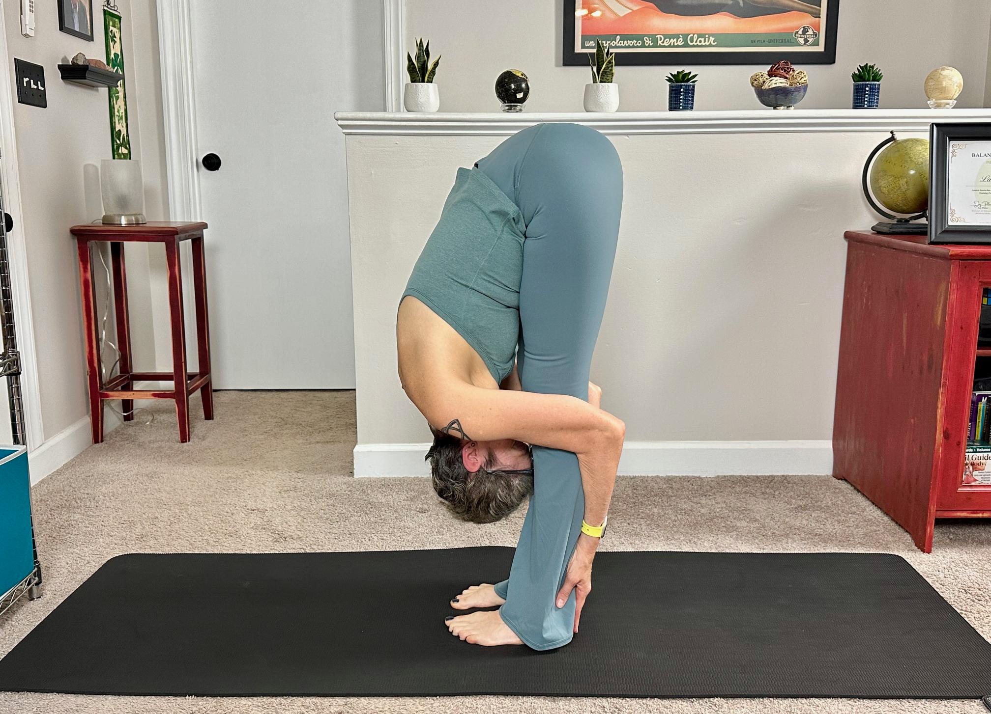 woman doing a standing forward fold on a yoga mat in a living room setting.