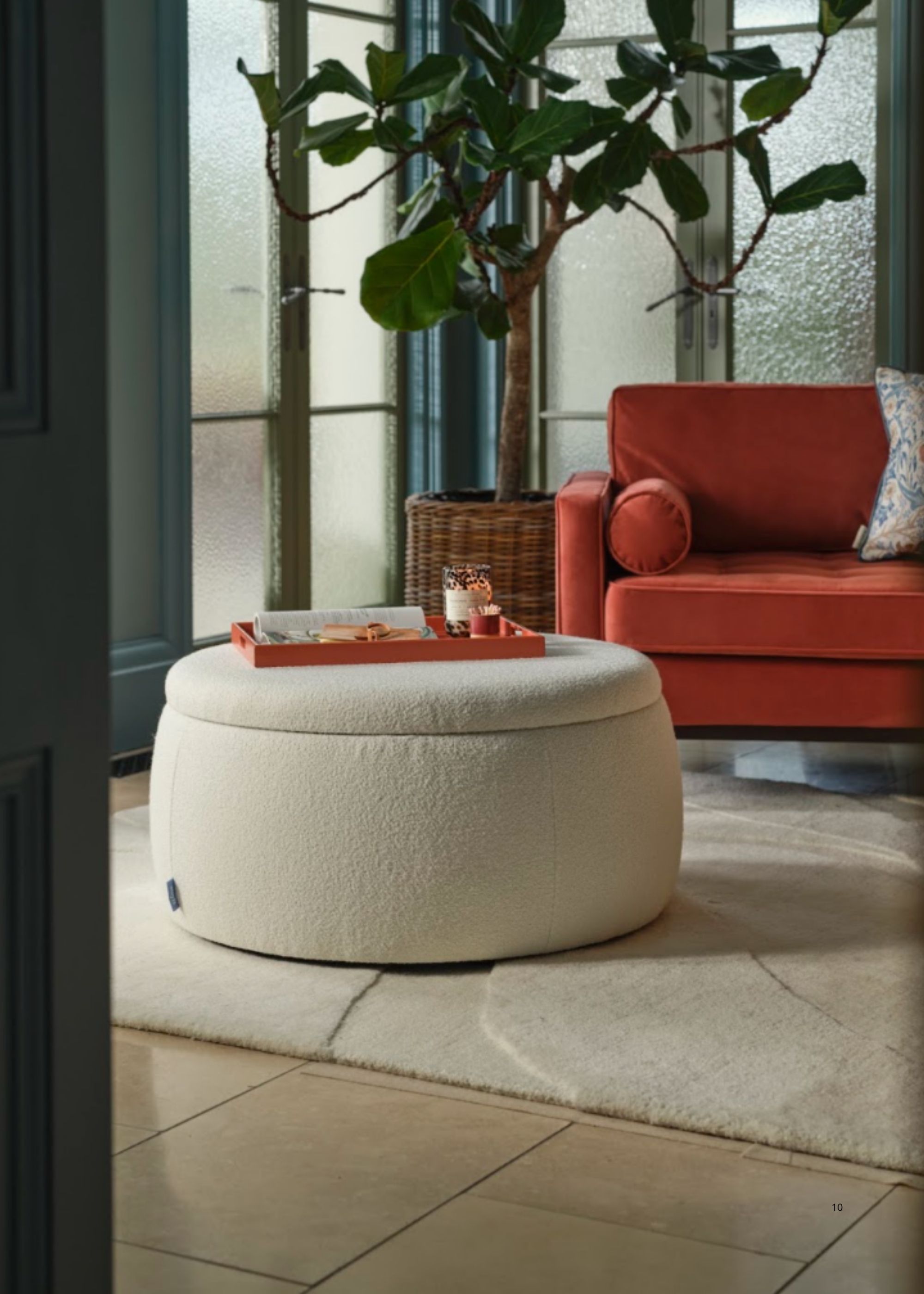 Image of a white boucle storage ottoman in a living room with a red sofa, a potted tree, a white rug, and a tray on top of the ottoman.