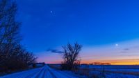 Bright star-like objects shine in the evening sky above a snowy road lined by trees on the left, as the glow of the setting sun lights up the horizon on the right of the image.