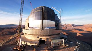 An enormous telescope dome is pictured in a desert during the day surrounded by cranes. Two huge silver sliding doors are visible, closed in the middle of the dome.