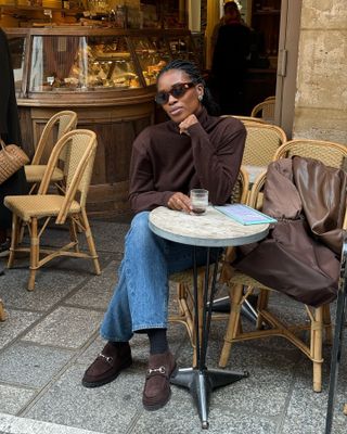 British fashion influencer Marilyn Nwawulor-Kazemaks sitting at a cafe in Paris wearing tort sunglasses, a brown turtleneck sweater, a straight-leg jeans, socks, and brown suede Gucci loafers.