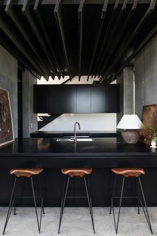 A modern kitchen with cement walls, black cabinetry and brown bar stalls