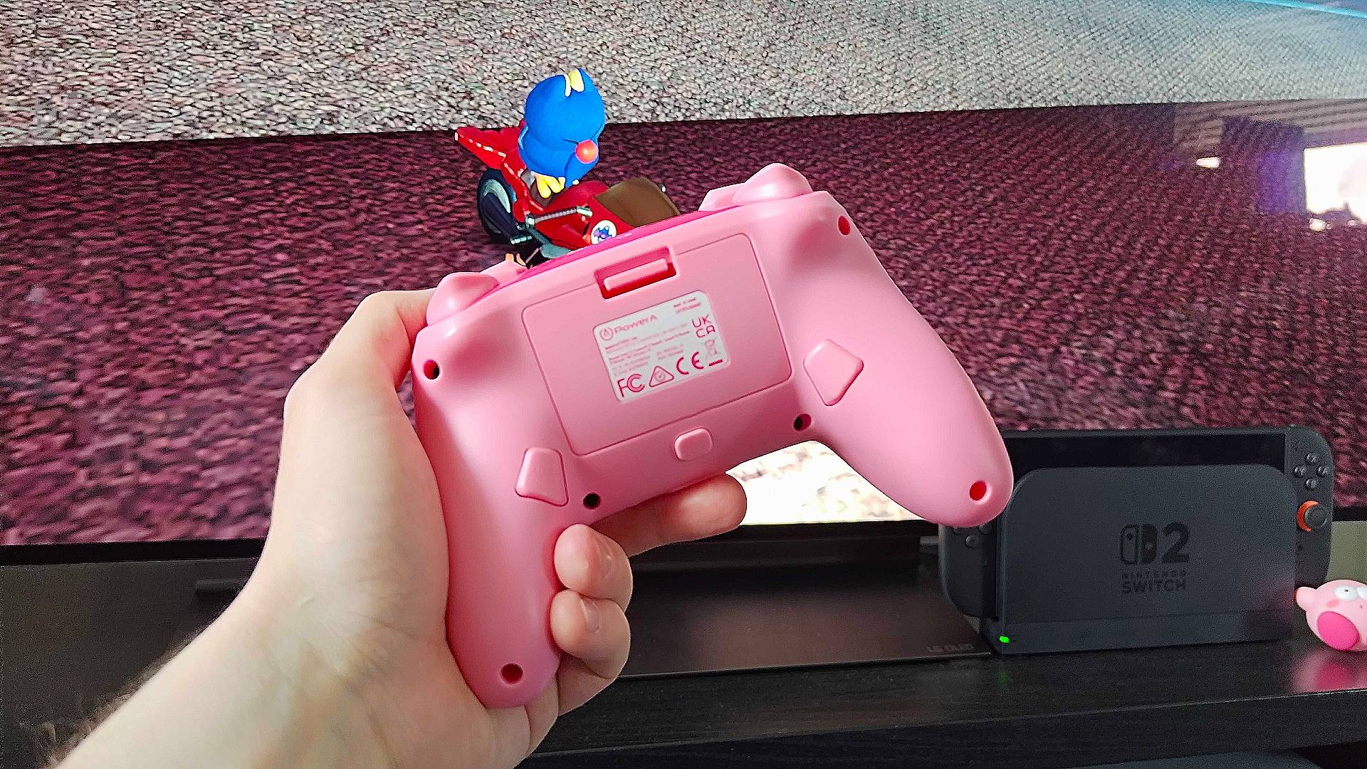 Hand holding PowerA Kirby Mouthful controller with Switch 2 and Mario Kart World running on TV in backdrop.