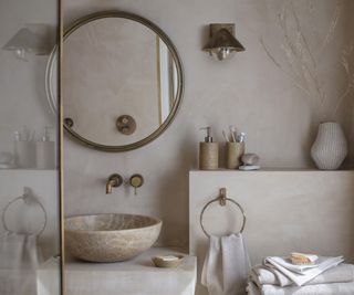 An earthy, minimalist bathroom featuring a natural stone basin atop a polished plaster countertop. Above the basin, a large round brass-framed mirror reflects light, complementing the warm tones of the wall-mounted brass taps.