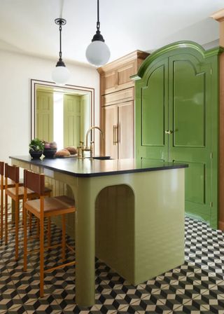 A sage green colored kitchen island with a dark countertop on top. Light pendants also hand above the island. Opposite the island is a large bright green kitchen cabinet. The flooring is a patterned tiling which adds a unique contrast to the space.