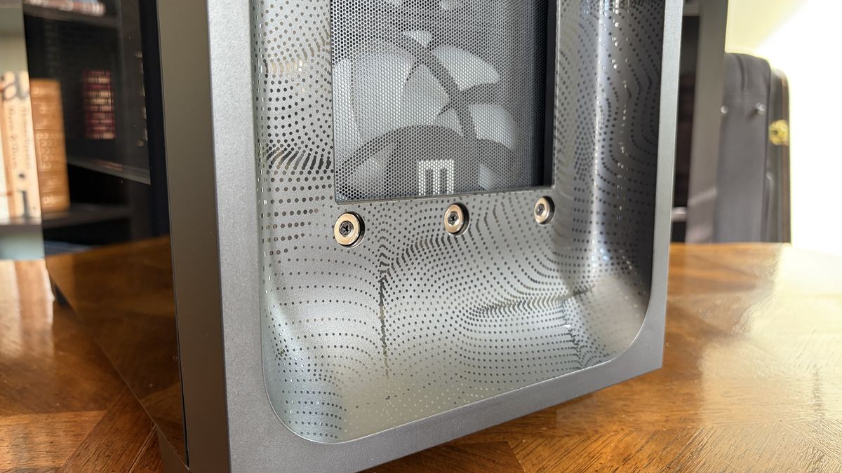 Close-up of a sleek MG-1 MK.II computer case with a mesh front panel featuring a dotted pattern and three visible screws. A fan with a logo is inside, set on a wooden table.