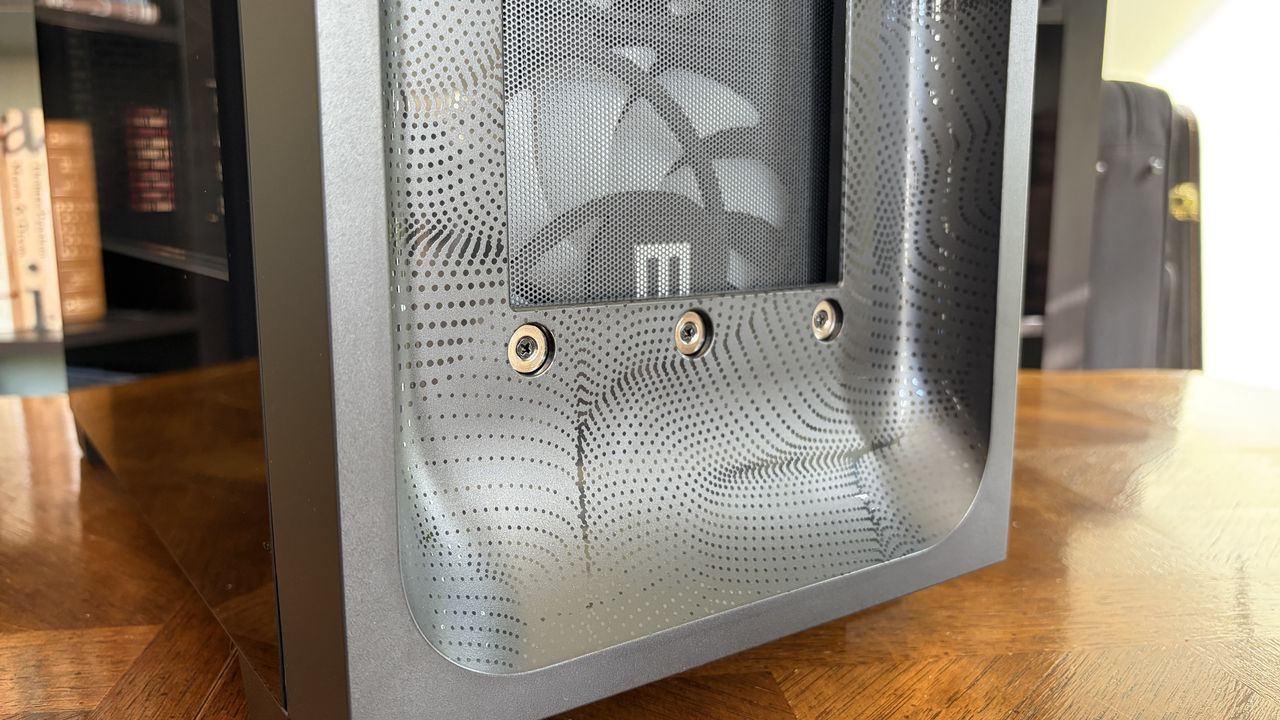 Close-up of a sleek MG-1 MK.II computer case with a mesh front panel featuring a dotted pattern and three visible screws. A fan with a logo is inside, set on a wooden table.