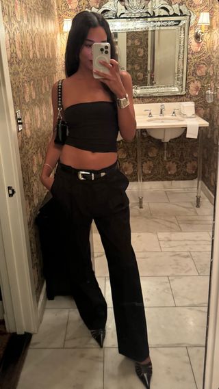 Laura Harrier taking a photo in the mirror in a bathroom wearing a black cropped tube top, Tiffany & Co. cuff bracelet, black trousers, black pumps, and a silver-and-black western belt.