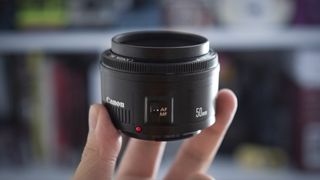 A closeup of the Canon EF 50mm f/1.8 STM lens being held in someone's fingertips