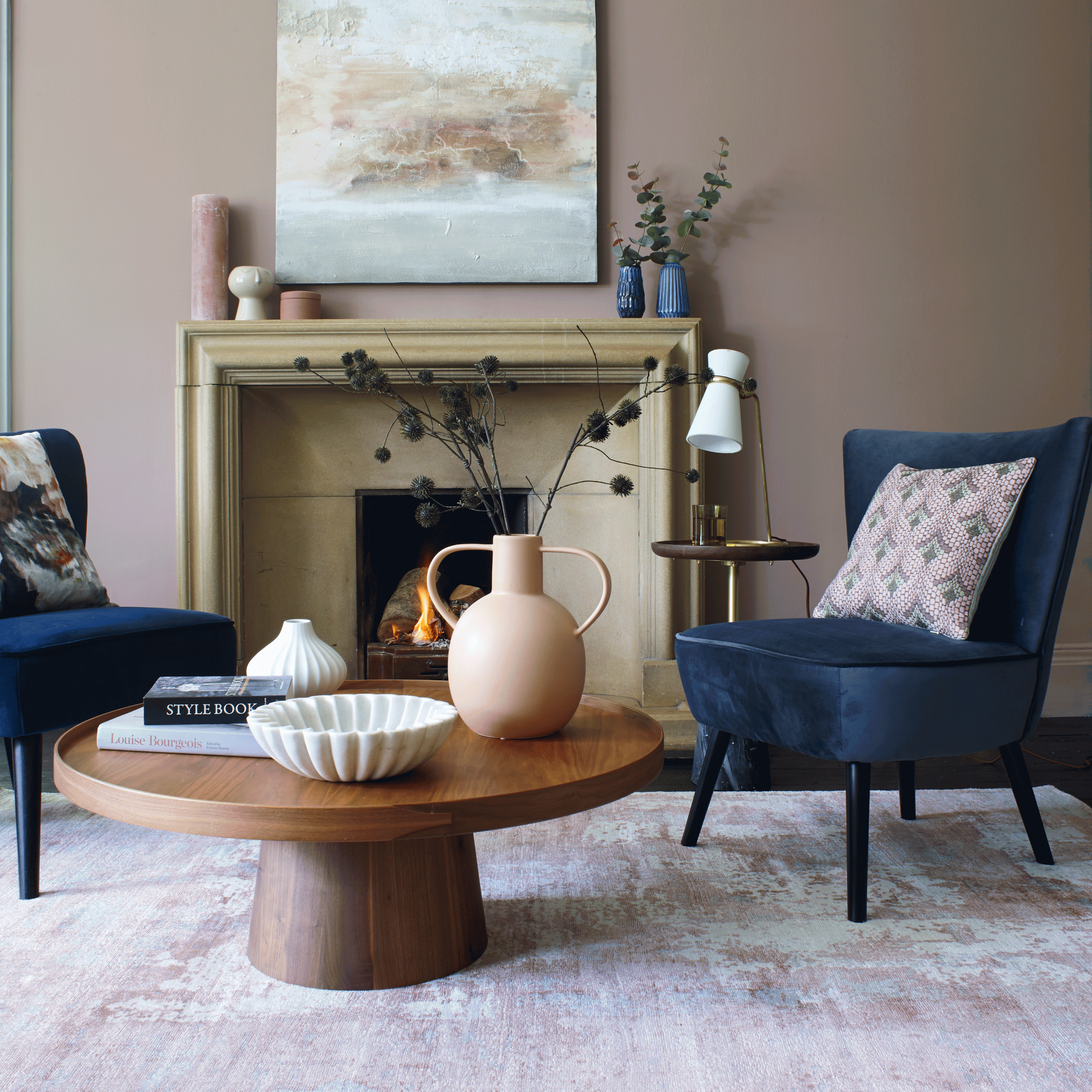 A natural wood coffee table with two blue upholstered chairs positioned in front of a lit fireplace.