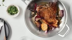 A large stainless steel frying pan with a carry handle on a white counter. A fried steak inside with red onion and a garlic bulb. Beside it is a small dish of chopped parsley, and a plate with a knife and fork.