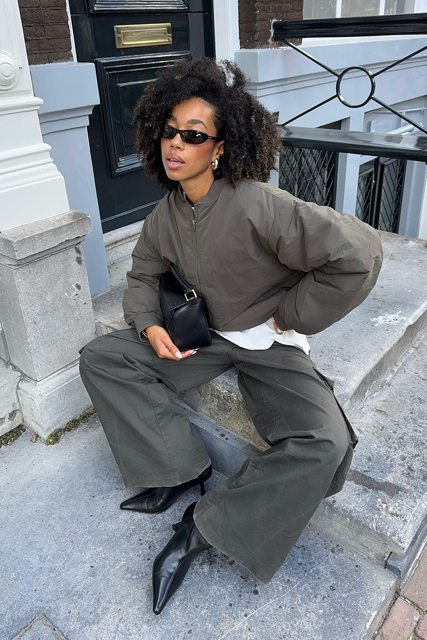 A woman wearing an olive bomber jacket, olive cargo pants, black shoes, black sunglasses and a black leather bag