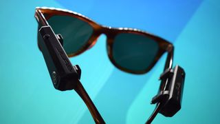 JLab JBuds Frames photograph showing devices on a pair of Ray-Ban sunglasses in front of a blue and teal background