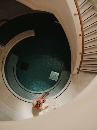 An aerial view of thermal baths in a luxury hotel shows a woman as she unwinds near a circular pool at the end of a stairway.