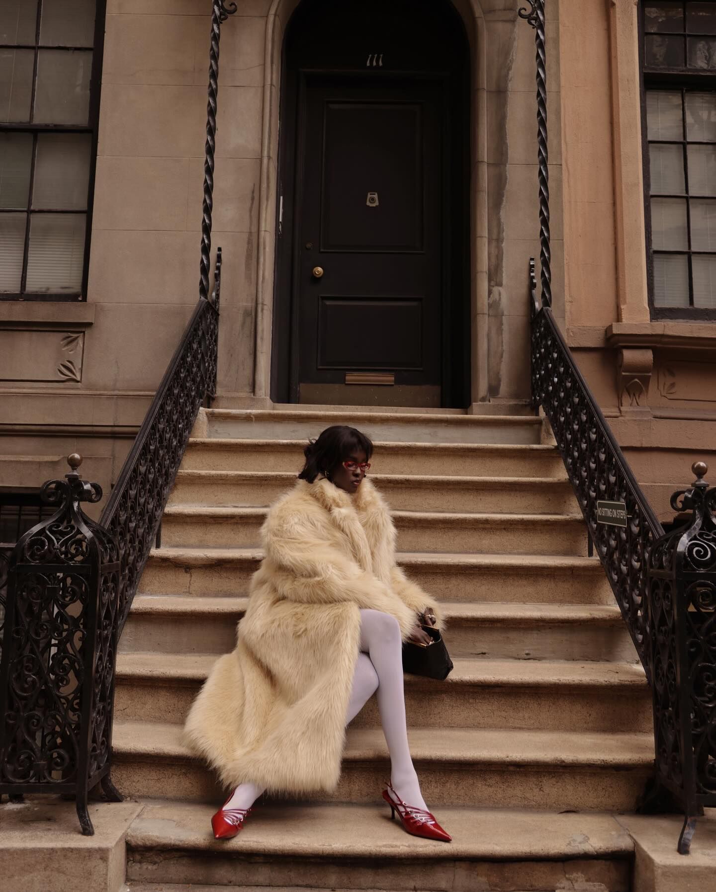 An image of @lefevrediary wearing a cream fur coat with white tights and red slingback heels.