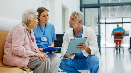 A doctor shows an older woman paperwork. She is discussing a procedure, advance directive, insurance or other matters. A nurse sits beside her.