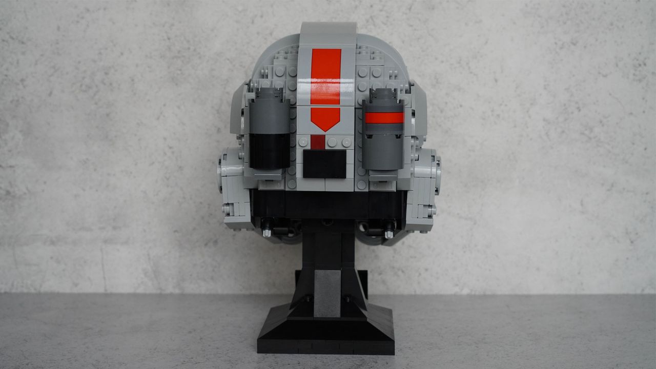A gray helmet with black eyes and frontal features also has splashes of red on the top and the bottom is on a black display stand with a name plaque in front of a gray background