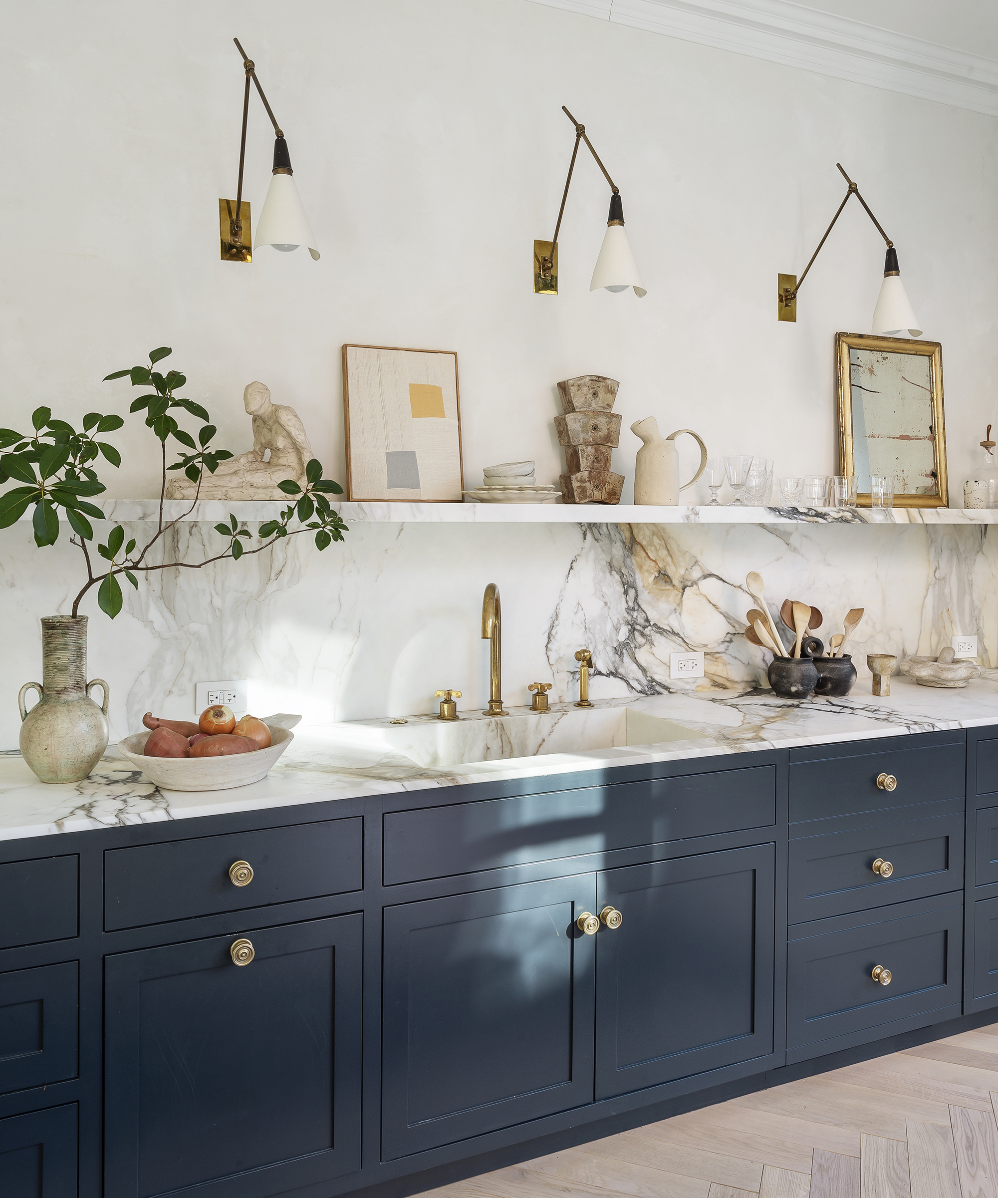 A kitchen with gold wall lights, marble surfaces and shelving and dark blue cabinetry.