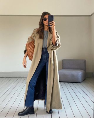 Marianne Smyth poses for a mirror selfie wearing a maxi trench coat, round metal-rim sunglasses, camel Dragon Diffusion woven tote bag, gray top, high-waisted straight-leg jeans, and flat black lace-up boots