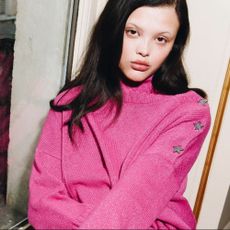 Woman with dark hair wearing bright pink jumper leaning against a window