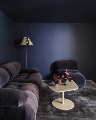 A moody cinema room. The walls are painted in a deep indigo paint color. There is a dark violet cloud sofa and matching accent chair arranged in the room. The rug is a grayish purple and there is a cream, metal coffee table between the seating.