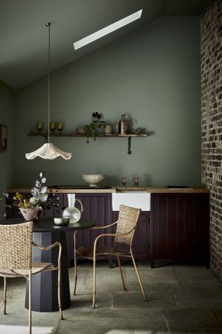 A moody kitchen palette with earthy green and burgundy, a brick wall and small dining table with a pretty low hung pendant above