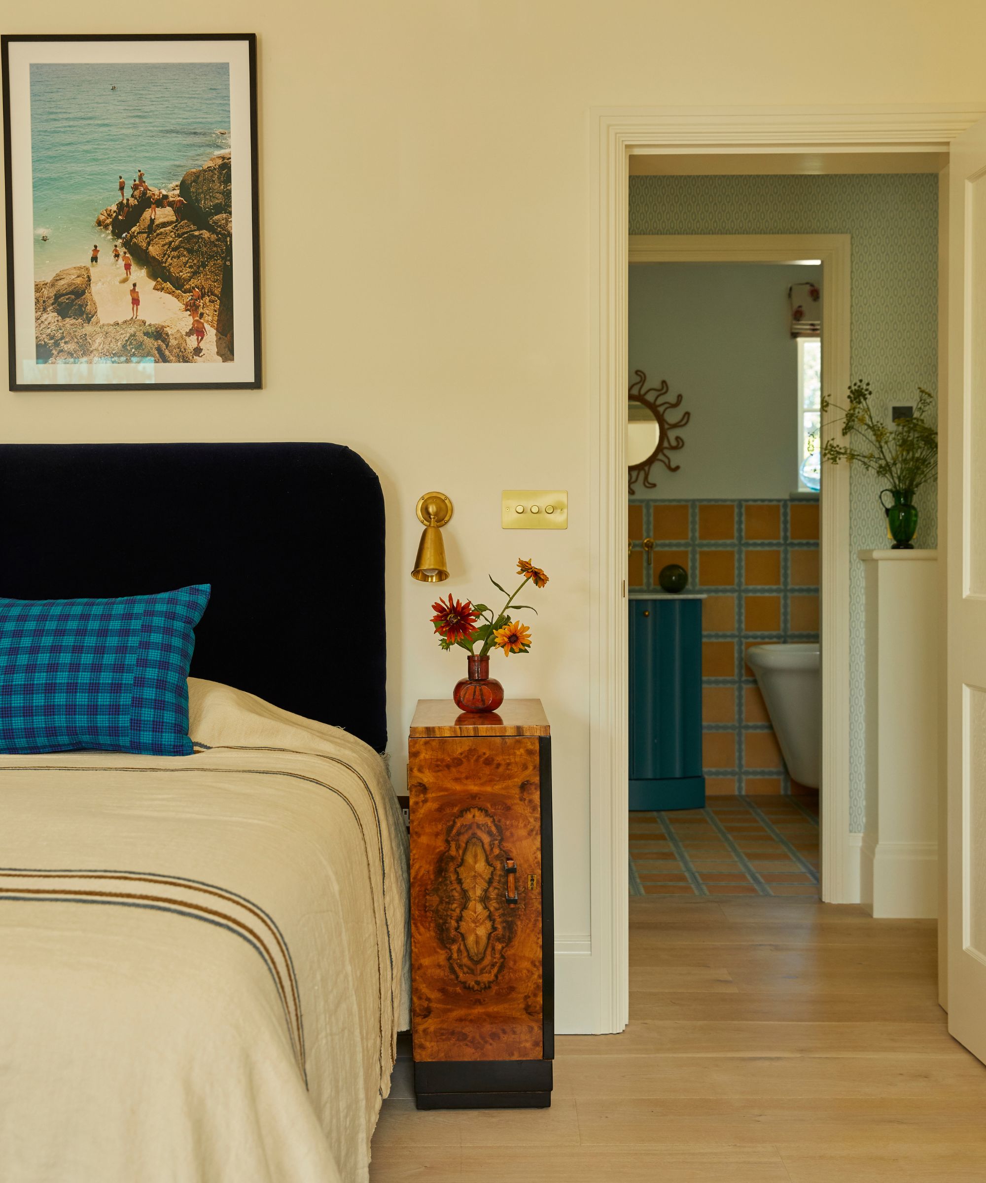A modern bedroom with warm white walls, creamy pale yellow bedding, a dark blue gingham cushion, and a coastal photograph on the wall. A doorway leading into a blue and yellow bathroom.