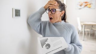 woman holding energy bill next to thermostat looking upset 