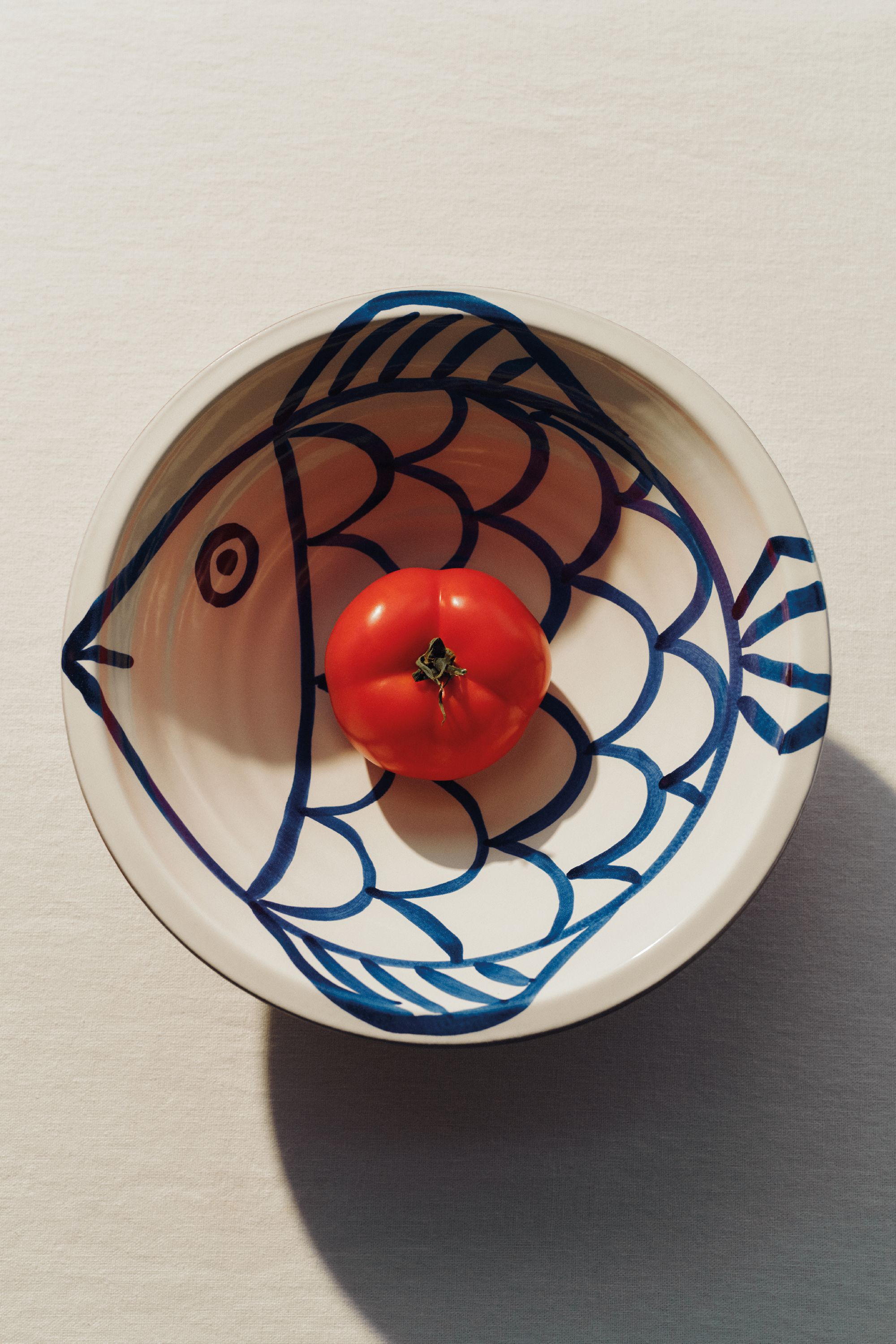 Image of a white bowl with a blue drawing of a fish on it. There is a single tomato in the bowl and the bowl is on a white tablecloth.