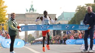 Eliud Kipchoge crosses the finish line of the Berlin Marathon, two people stand either side holding the finish line tape, the Brandenburg Gate in the background