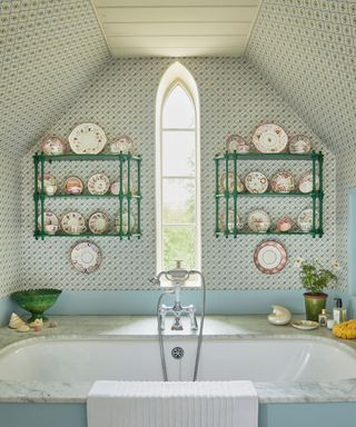 blue bathroom with blue, green and white patterned wallpaper on walls and part of the ceiling, two matching green shelves decorated with plates either side of the unique arch window, built-in bath with marble top.