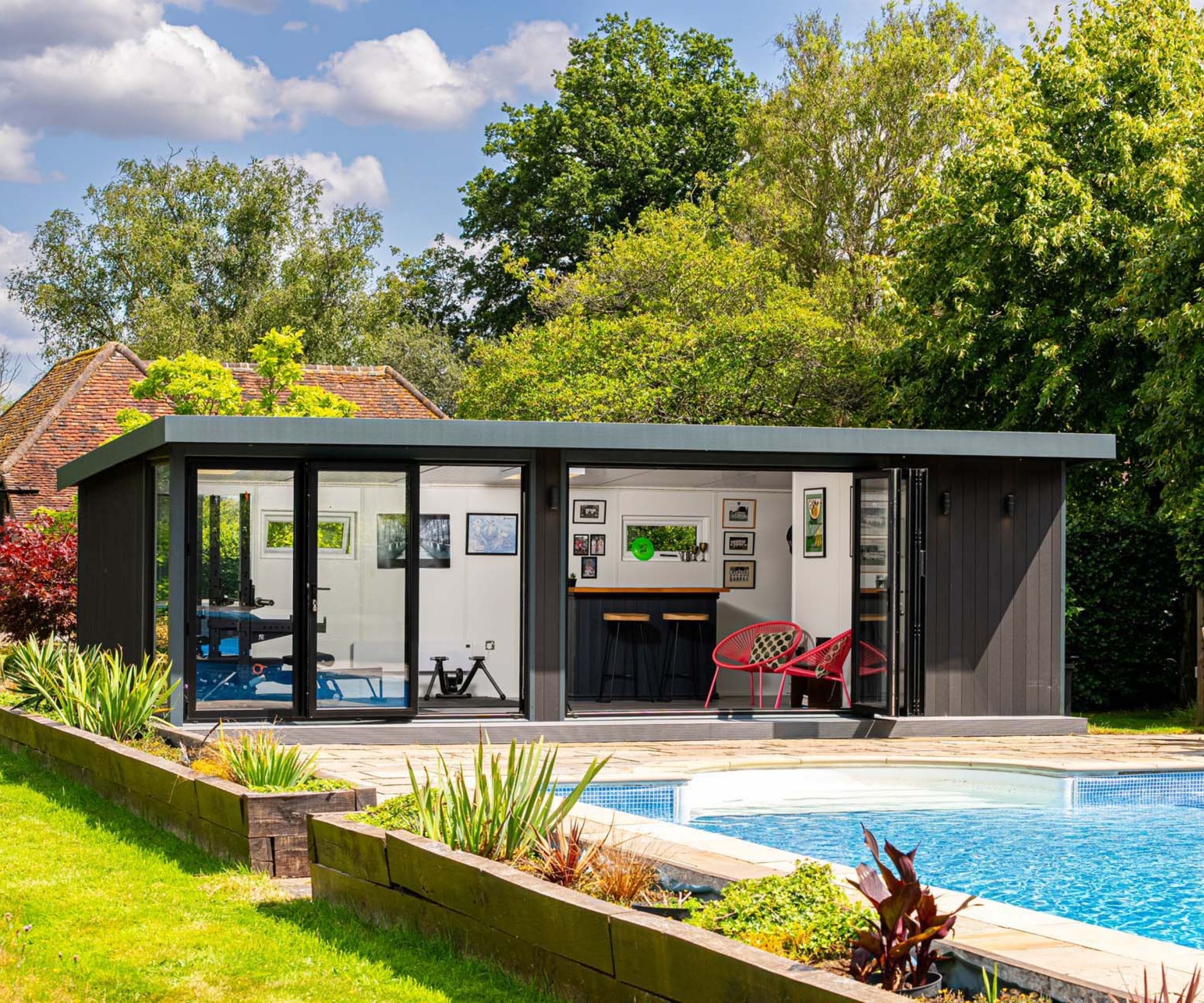 A dark grey flat-roof garden room with full-width bifold doors open to reveal a gym on one side and a bar with red chairs on the other, beside a mosaic-tiled swimming pool.