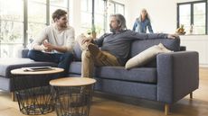 A young man smiles as he talks with his father on the sofa while his mother smiles in the background.